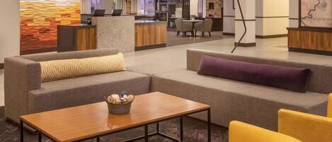 A modern hotel lobby with a sectional sofa, yellow armchairs, a wooden coffee table, and a reception desk.