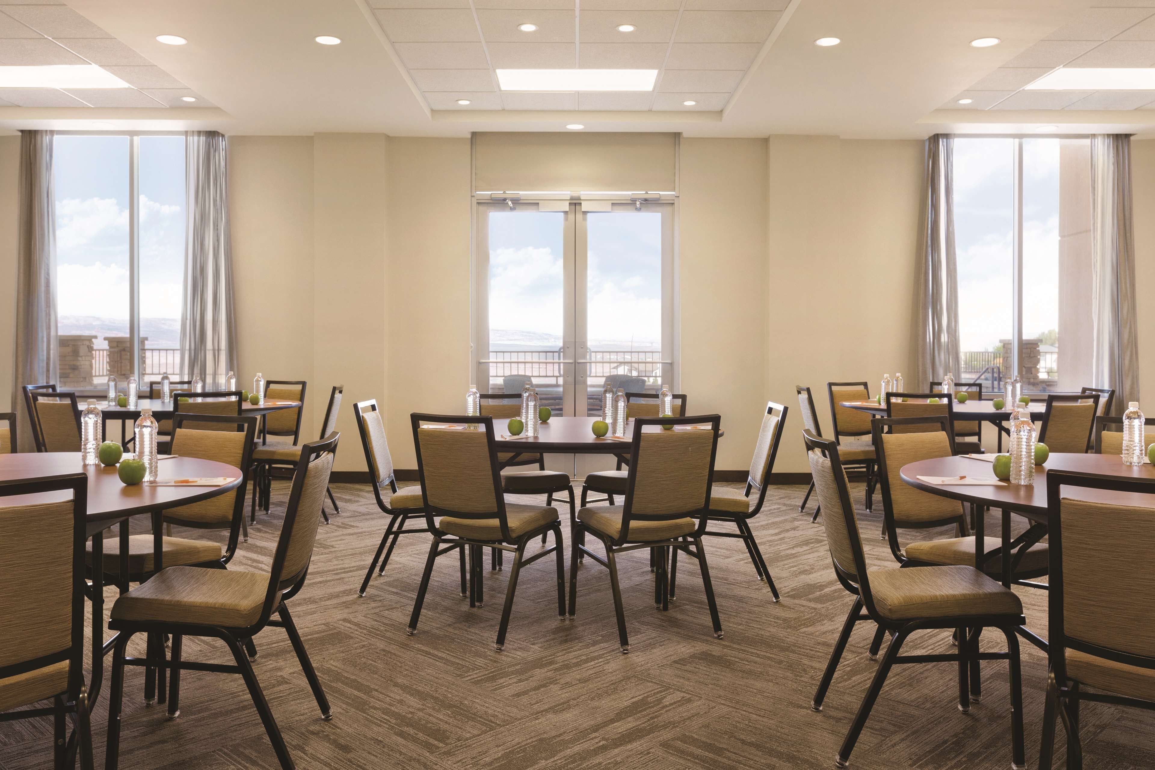 A hotel conference room with multiple tables and chairs, large windows with curtains, and a view of the cityscape.