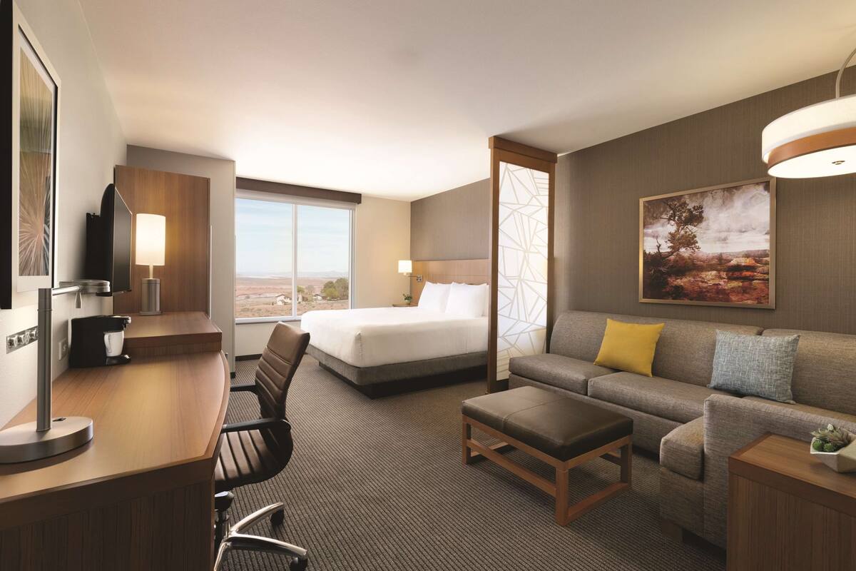 A hotel room with a bed, a sofa, a desk, and a chair. There is a large window providing a view of the outdoors.