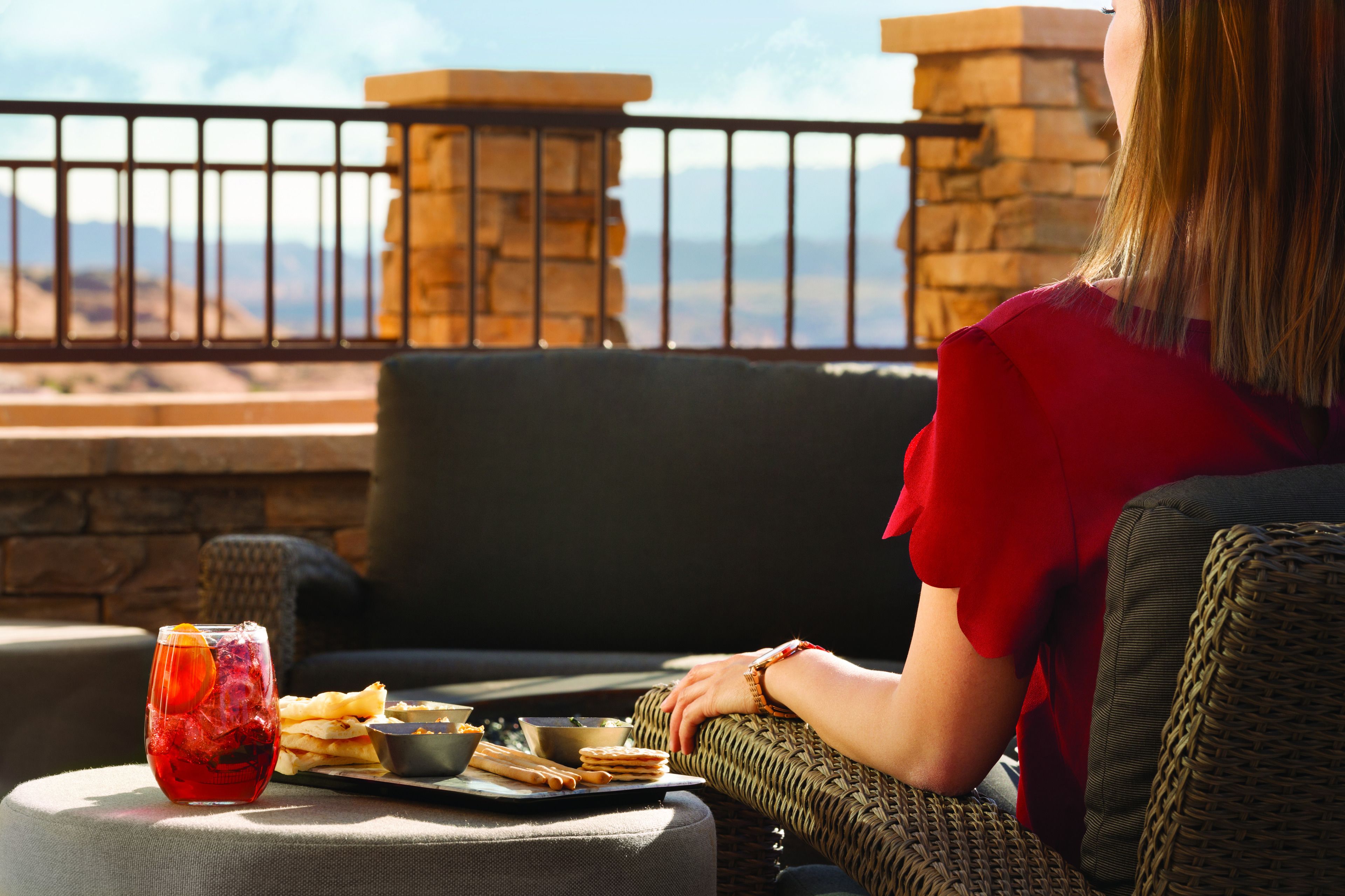A person seated on a wicker sofa with a table in front of them, containing a drink, snacks, and a tray.