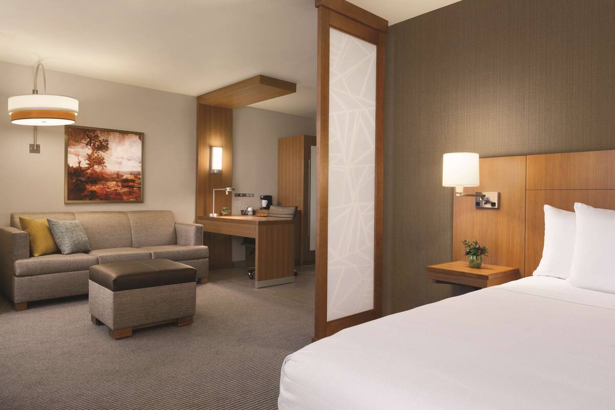 A hotel room with a bed, a sofa, a desk, and a chair. There is a painting on the wall and a lamp on the desk.