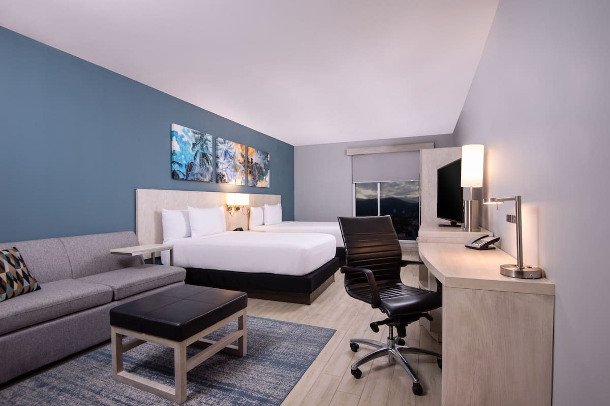 Ein modernes Hotelzimmer mit einem großen Bett, einem Schreibtisch mit Lampe, einem Sessel, einer Couch und Bildern an der Wand.