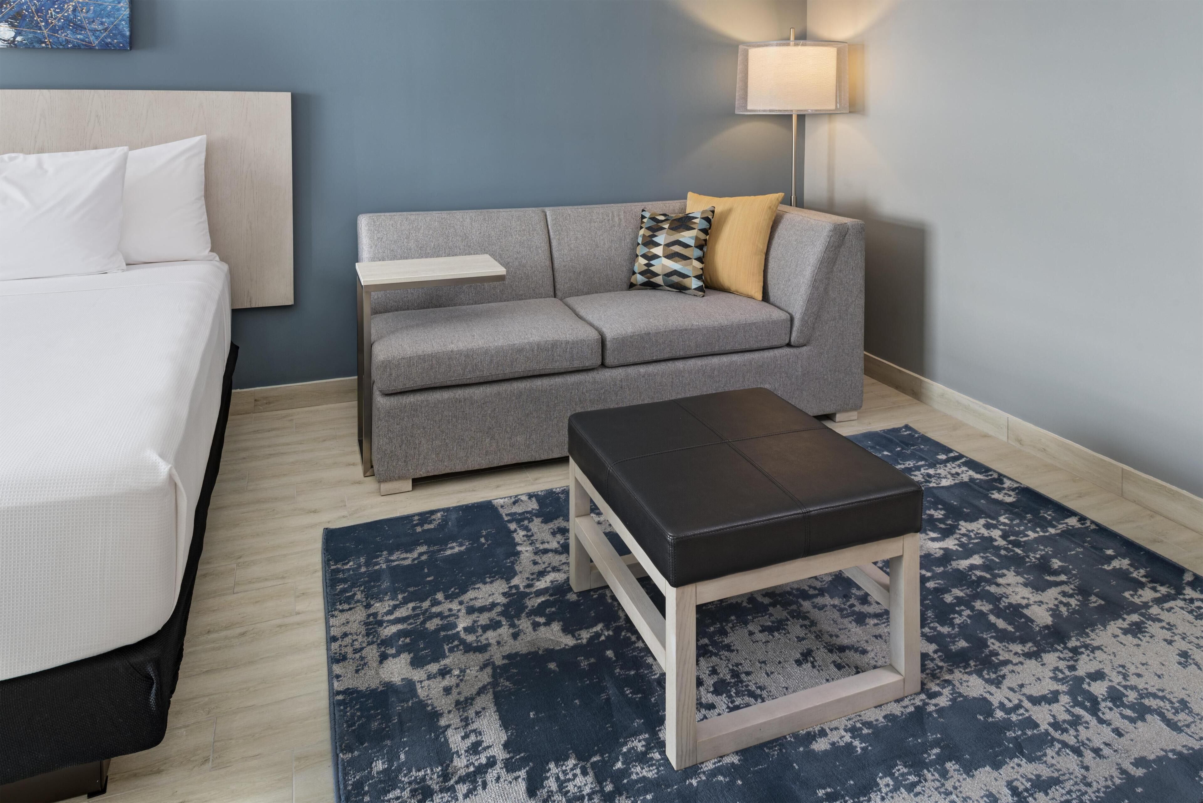 Una habitación de hotel con una cama, un sofá gris con cojines, una mesita, un otoman negro y una alfombra azul estampada.