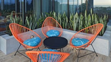 Un espacio al aire libre moderno con sillas naranjas y una mesa negra, rodeado de plantas verdes grandes.