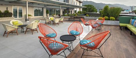 Un patio en la azotea con sillas de mimbre y metal, un sofá verde y una terraza con cierre de vidrio.