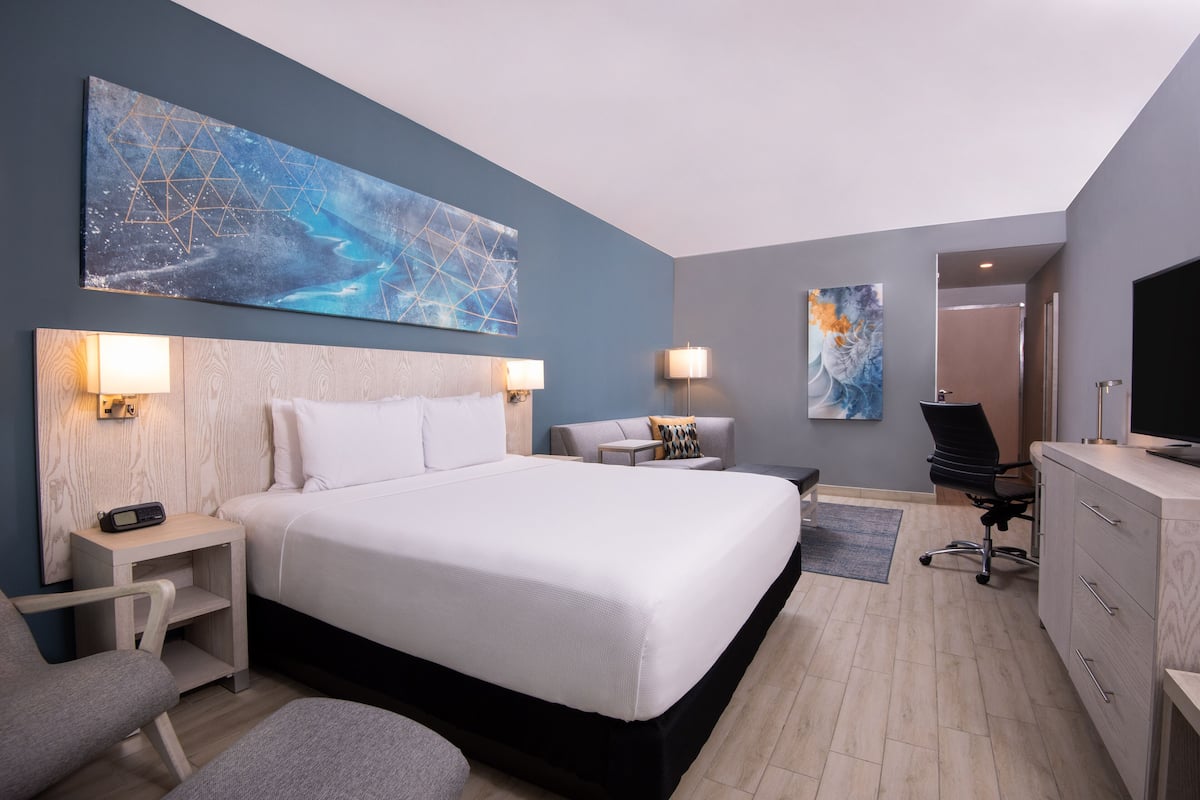 A modern hotel room with a large bed, bedside lamps, a desk, a chair, and artwork on the walls.