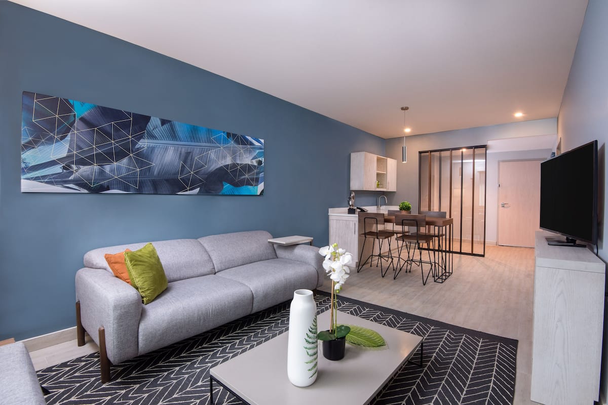 Ein modernes Wohnzimmer mit grauem Sofa, Couchtisch mit Vase und Orchidee, großem abstrakten Gemälde an der Wand und Küchenbereich im Hintergrund.