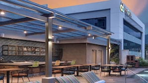 Un espace extérieur aménagé avec des fauteuils et des tables, un bar avec un mur en briques, et un bâtiment sur lequel on peut lire l’inscription « AGORA ».