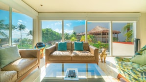 A spacious living room with a large window offering a view of a tropical landscape, a leather sofa, and a glass coffee table with remote controls. - Castillo de Arena by NAS (Playa del Carmen)