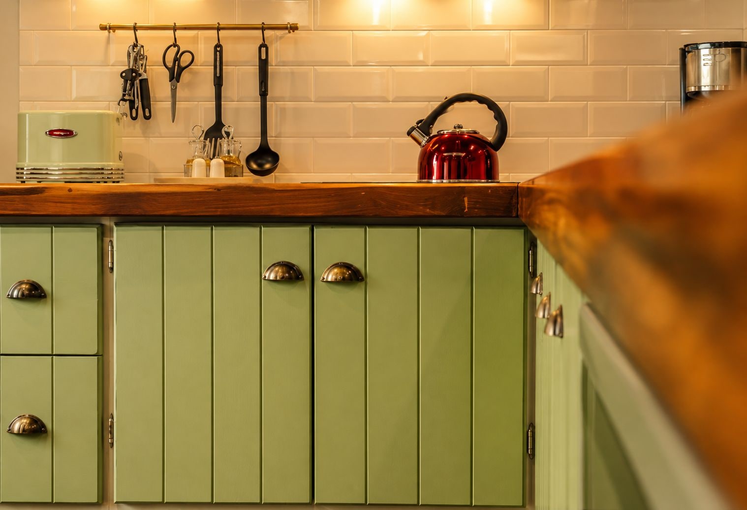 A kitchen with green cabinets, a red kettle, and various utensils hanging on the wall.