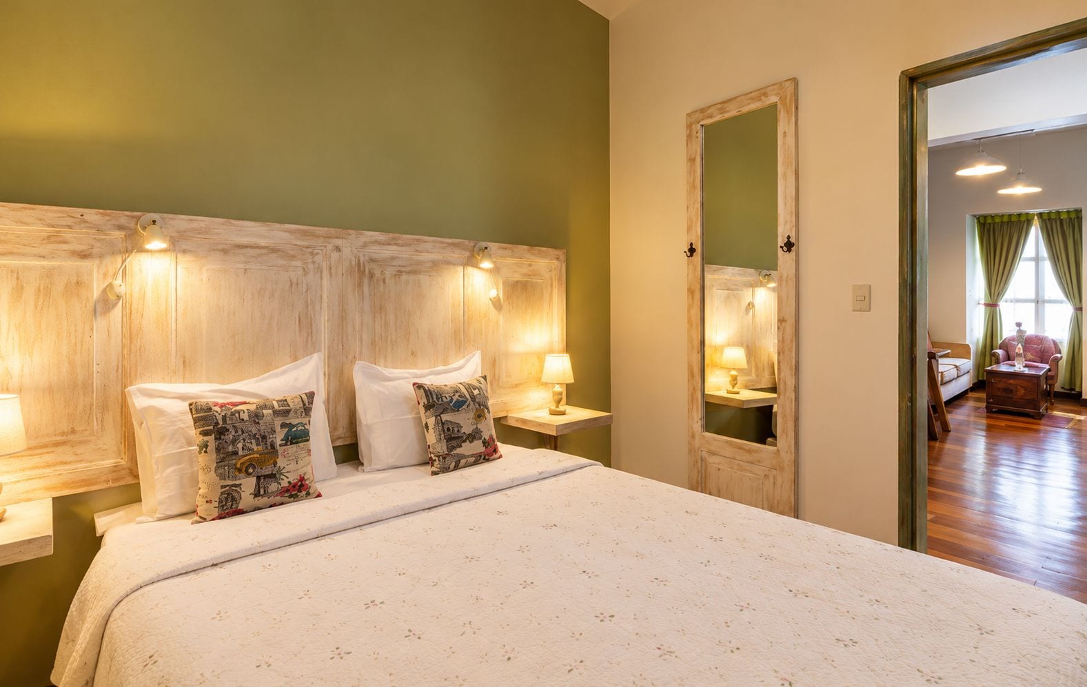 A hotel room with a large bed, bedside lamps, a mirror, and a view of the adjacent room.