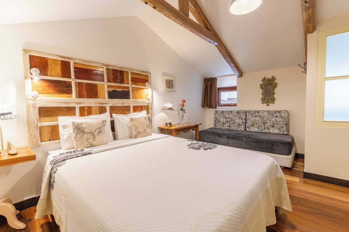 A bedroom with a large bed, a wooden headboard, a small bedside table, a nightstand, a bench, and a window with curtains.
