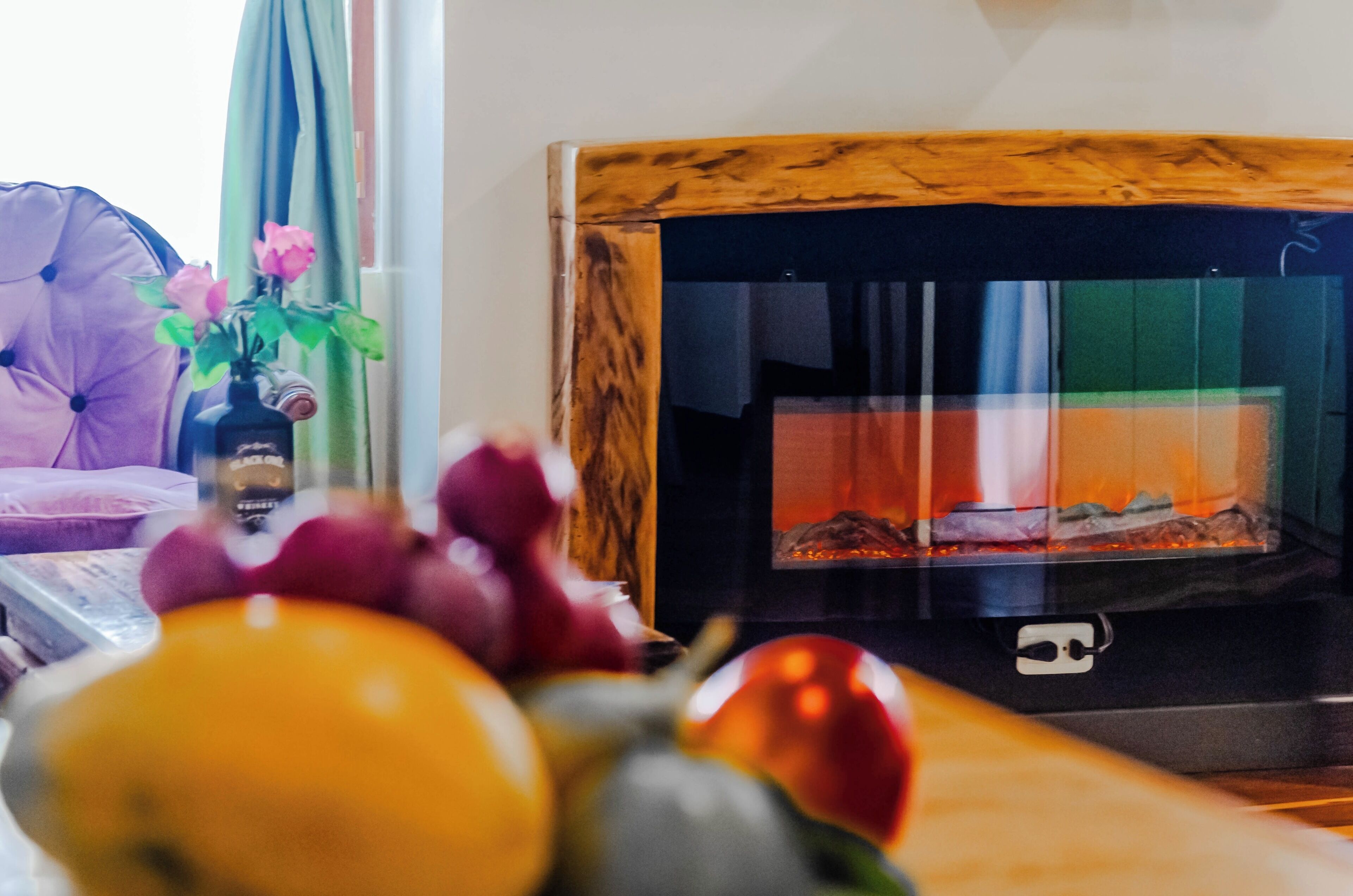 Een woonkamer met een open haard, een bank, een vaas met bloemen en een schaal met fruit.