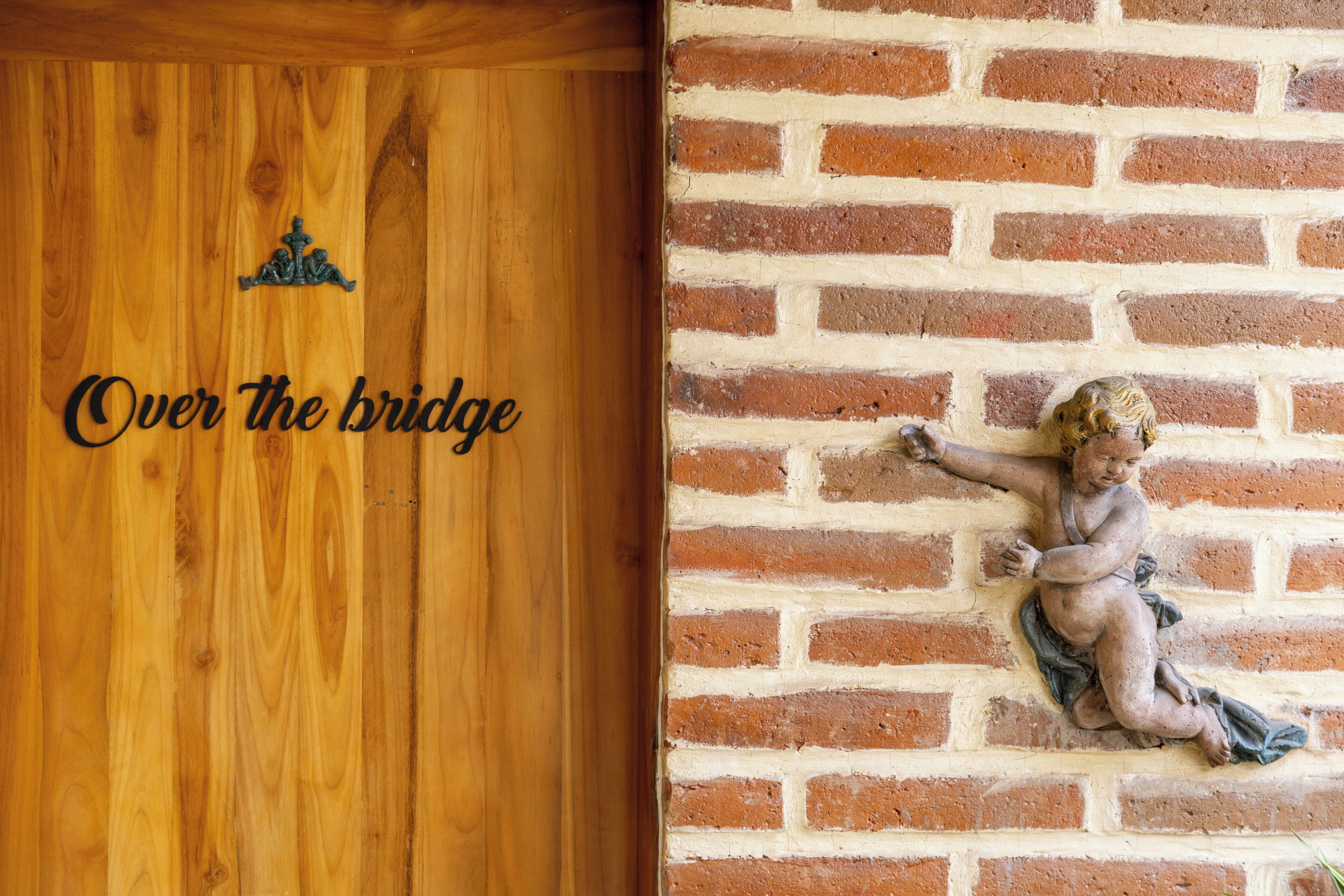 Una puerta de madera con la inscripción “Over the bridge” y un muro de ladrillo con una estatua de un ángel.