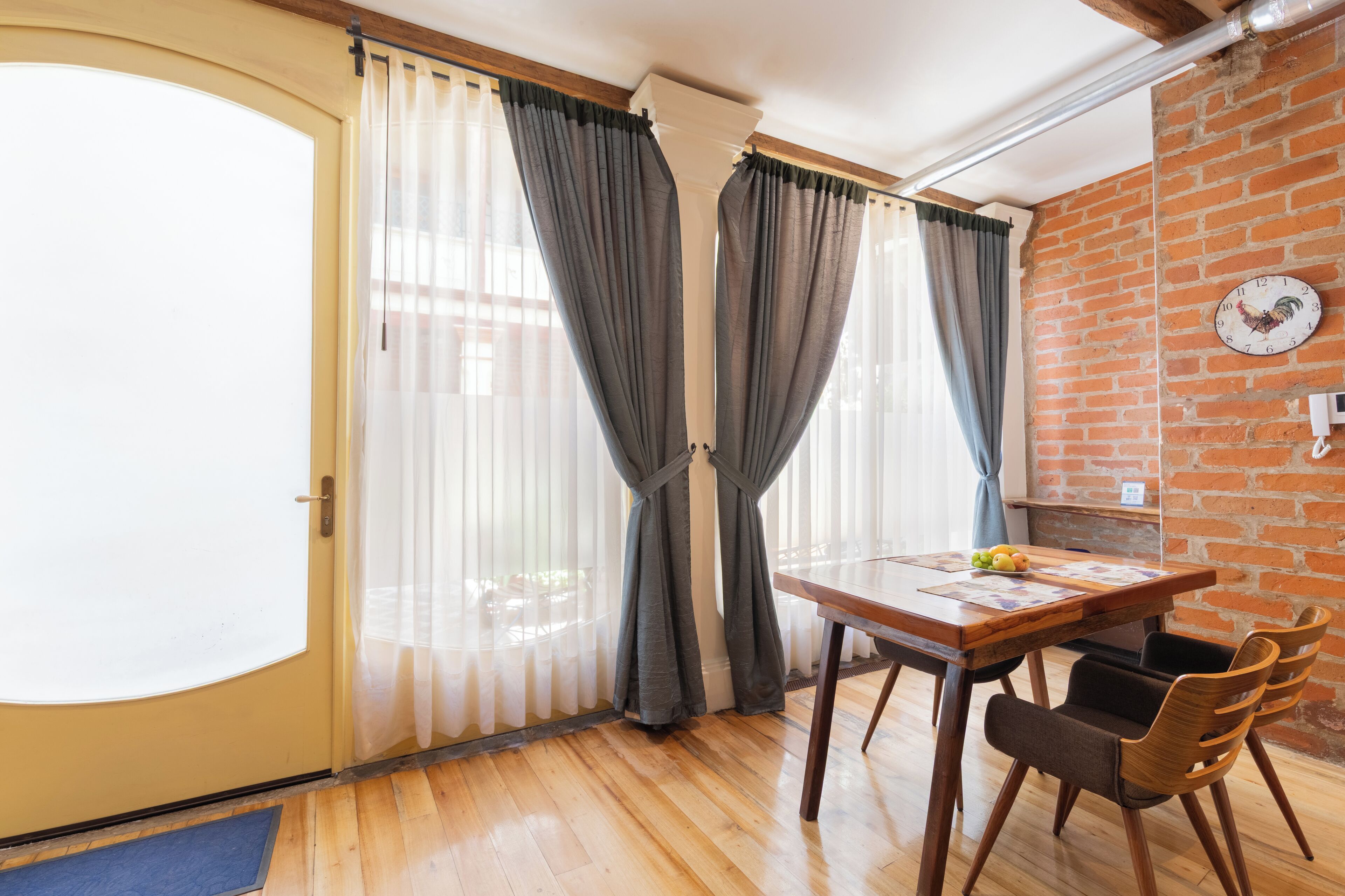 A room with a wooden floor, a dining table with chairs, a clock on the wall, and a door with curtains.