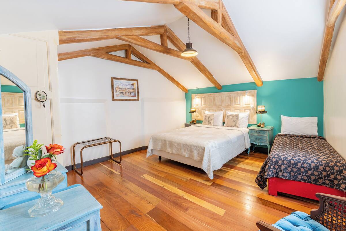 A bedroom with a wooden ceiling, a bed with a patterned bedspread, a bedside table with a lamp, and a dresser with a vase of flowers.