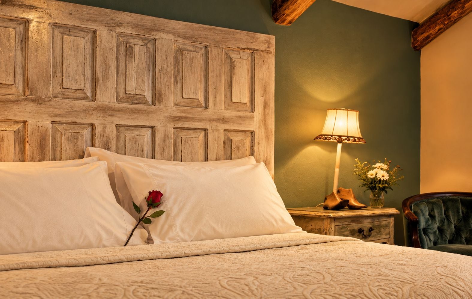 Una cama con sábanas blancas, cabecera de madera, lámpara de noche, un florero con flores y una silla.