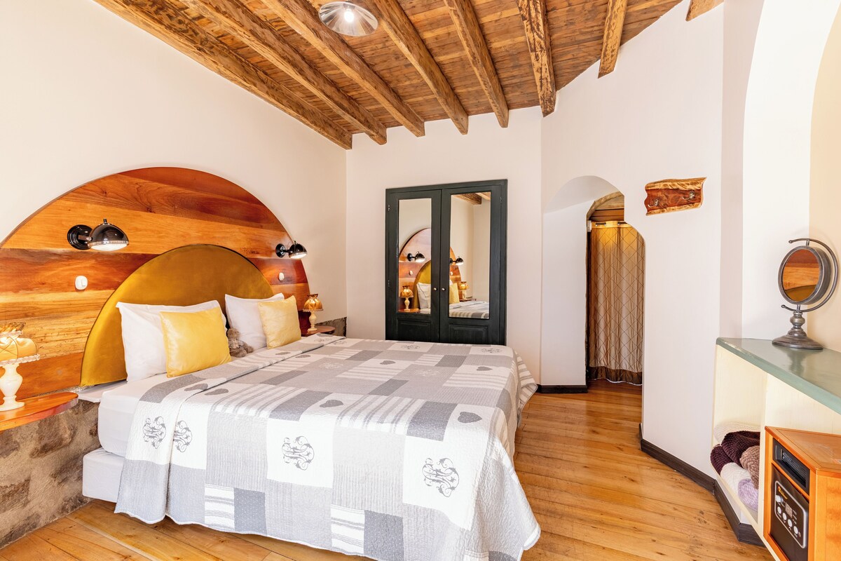 A bedroom with a wooden ceiling, a bed with a patterned bedspread, a mirror, and a small table with a lamp.