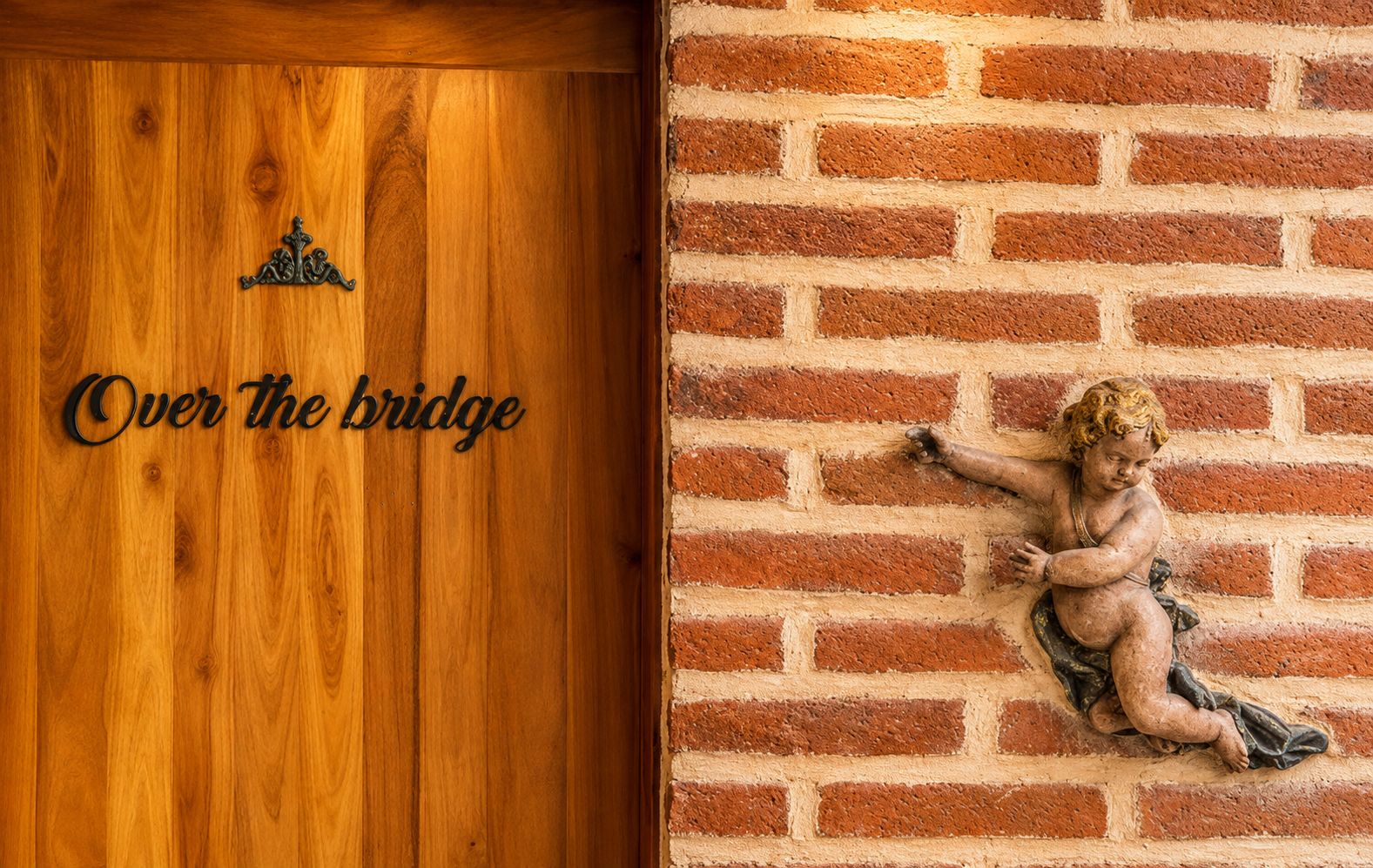 A wooden door with the inscription 'Over the bridge' and a brick wall with a statue of a cherub.