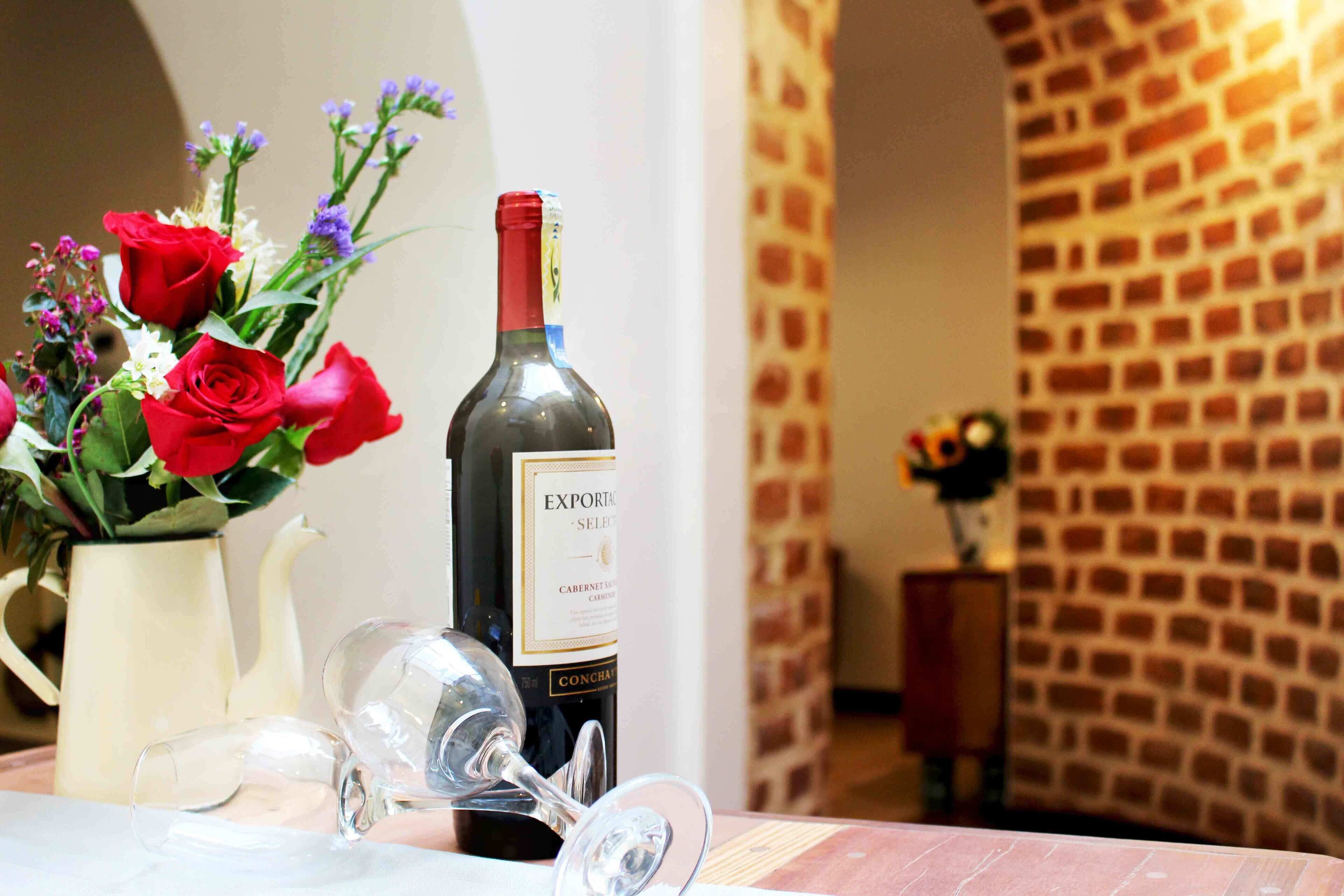 Une bouteille de vin et des verres sur une table, avec un vase de roses rouges et d’autres fleurs.