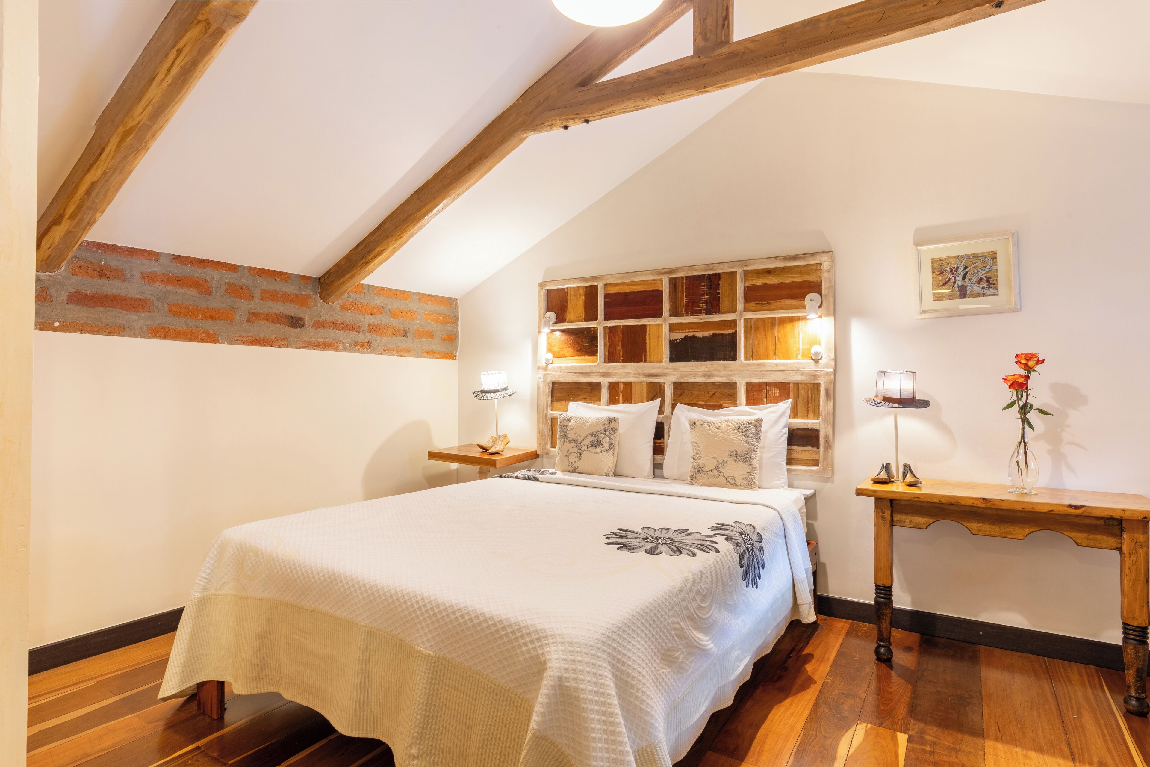 A bedroom with a wooden ceiling, a brick wall, a bed with a white bedspread, a bedside table with a lamp, and a small table with a vase of flowers.