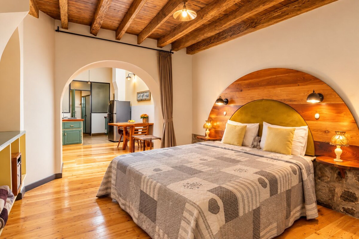 A bedroom with a wooden headboard, a bed with patterned bedding, a dining area with a table and chairs, and a kitchen in the background.
