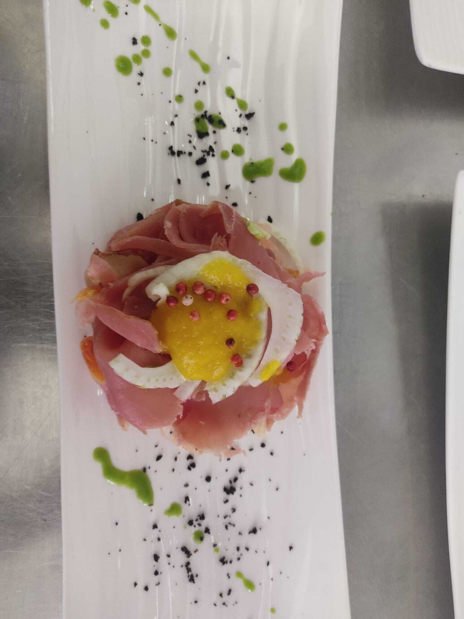 A white plate with a dish of prosciutto, egg, and a yellow sauce, garnished with green and black specks.