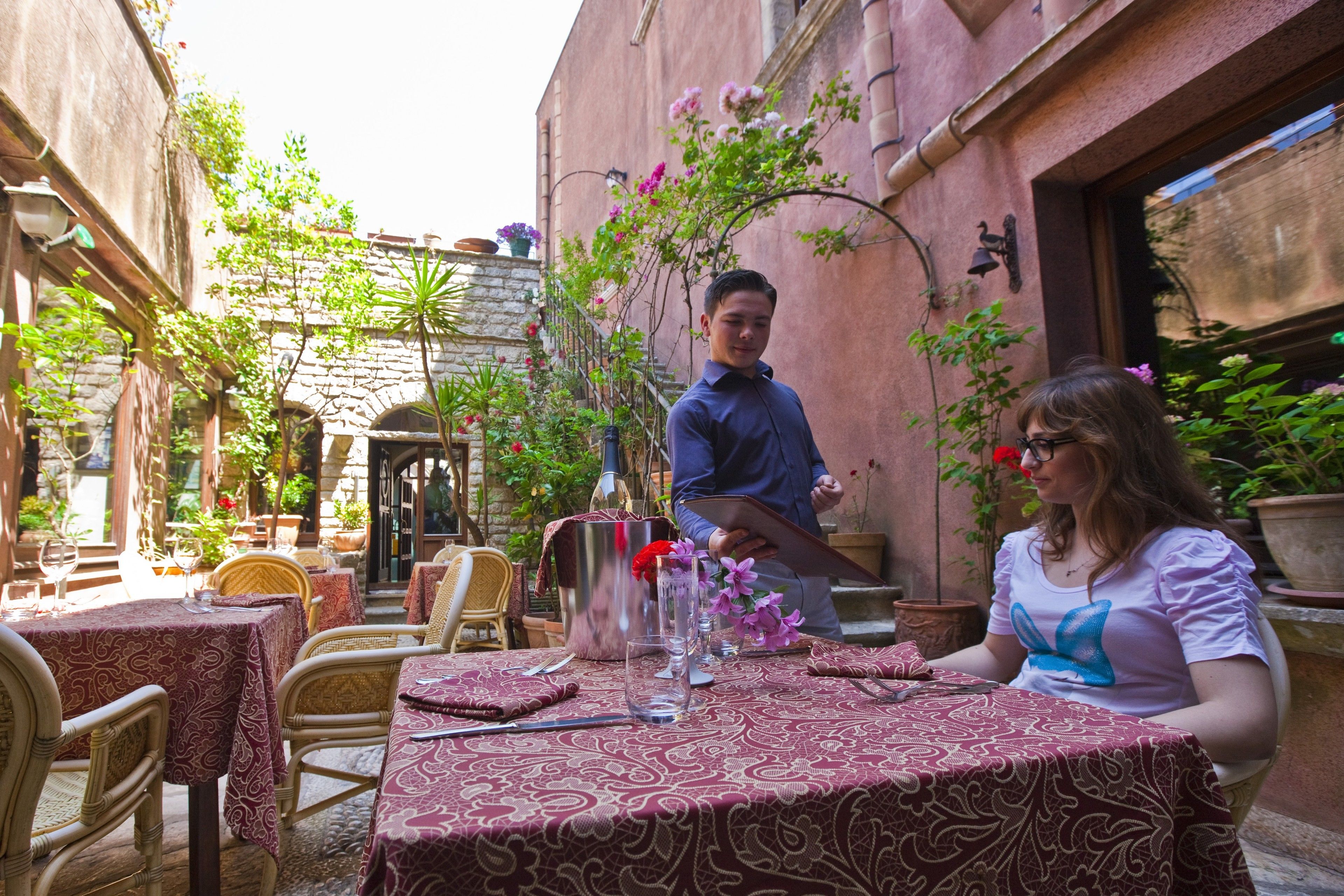 Un área de comedor al aire libre con mesas cubiertas de manteles estampados, rodeada de plantas en macetas y un muro de piedra.