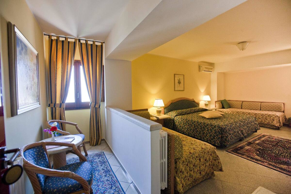 Habitación de hotel con dos camas, una mesita con una silla, un radiador y una ventana con cortinas.