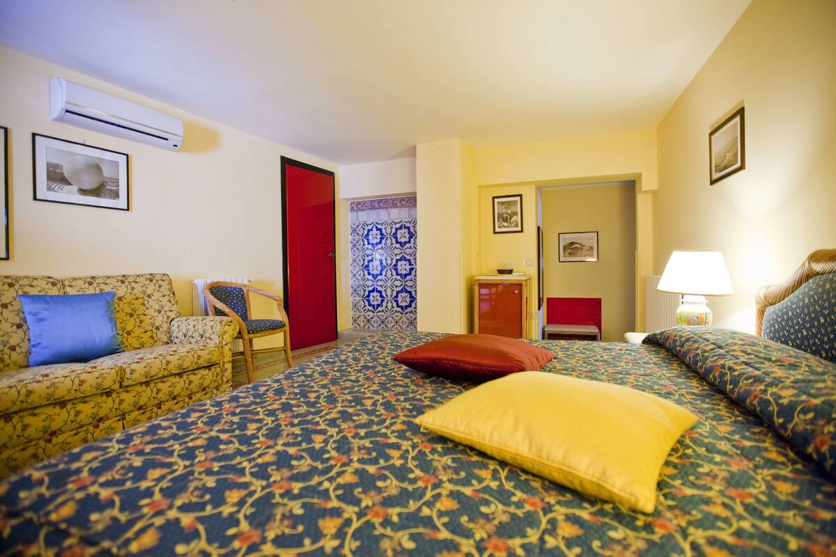 Habitación de hotel con alfombra estampada, un sofá, una cama con almohada amarilla, una silla, una puerta roja y un aire acondicionado instalado en la pared.