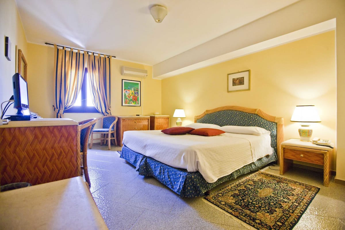 Una habitación de hotel con cama, escritorio, silla, mesita de noche, lámpara y alfombra.