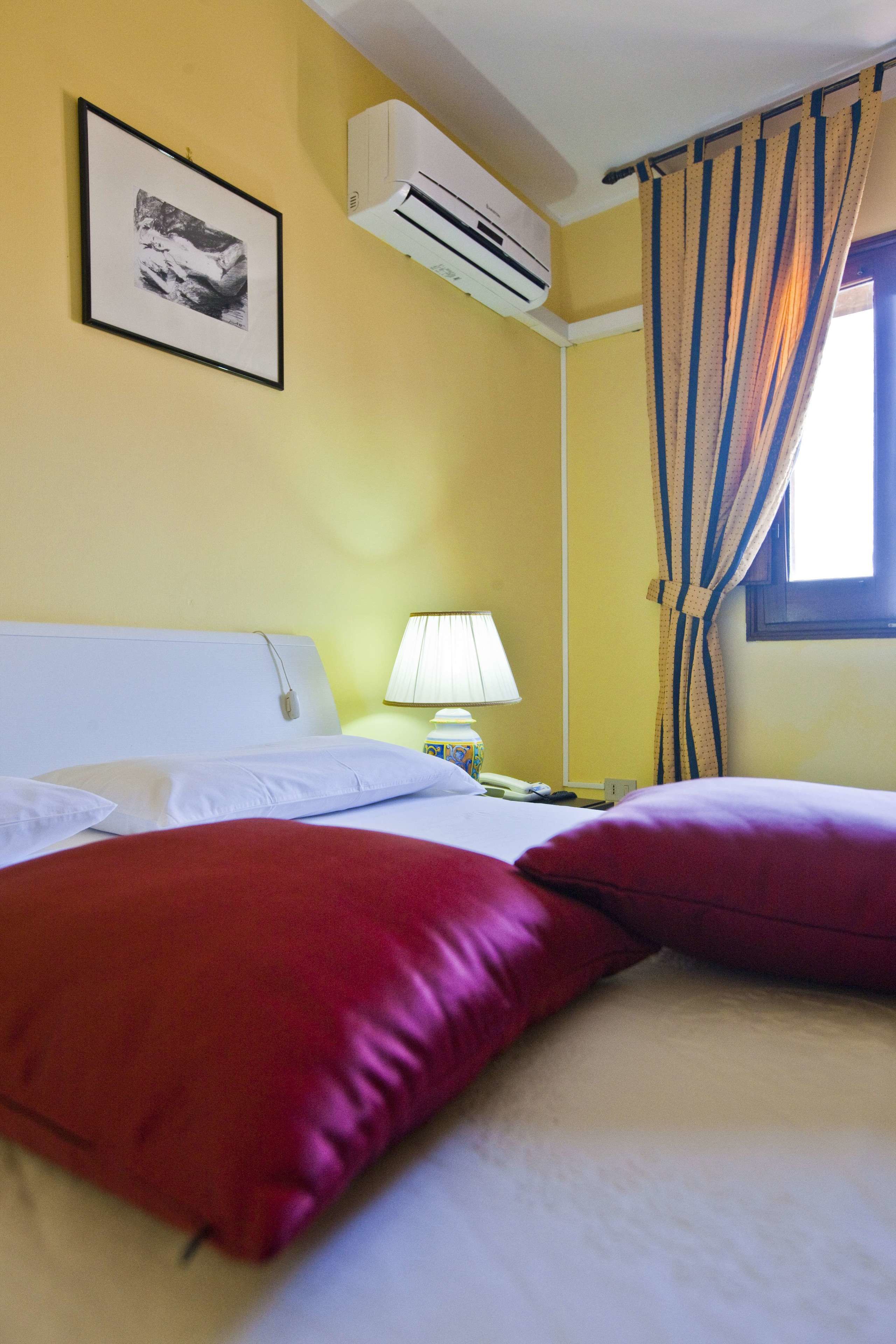 Habitación de hotel con cama, almohadas, mesita de noche con lámpara, aire acondicionado, un cuadro en la pared y una ventana con cortinas.
