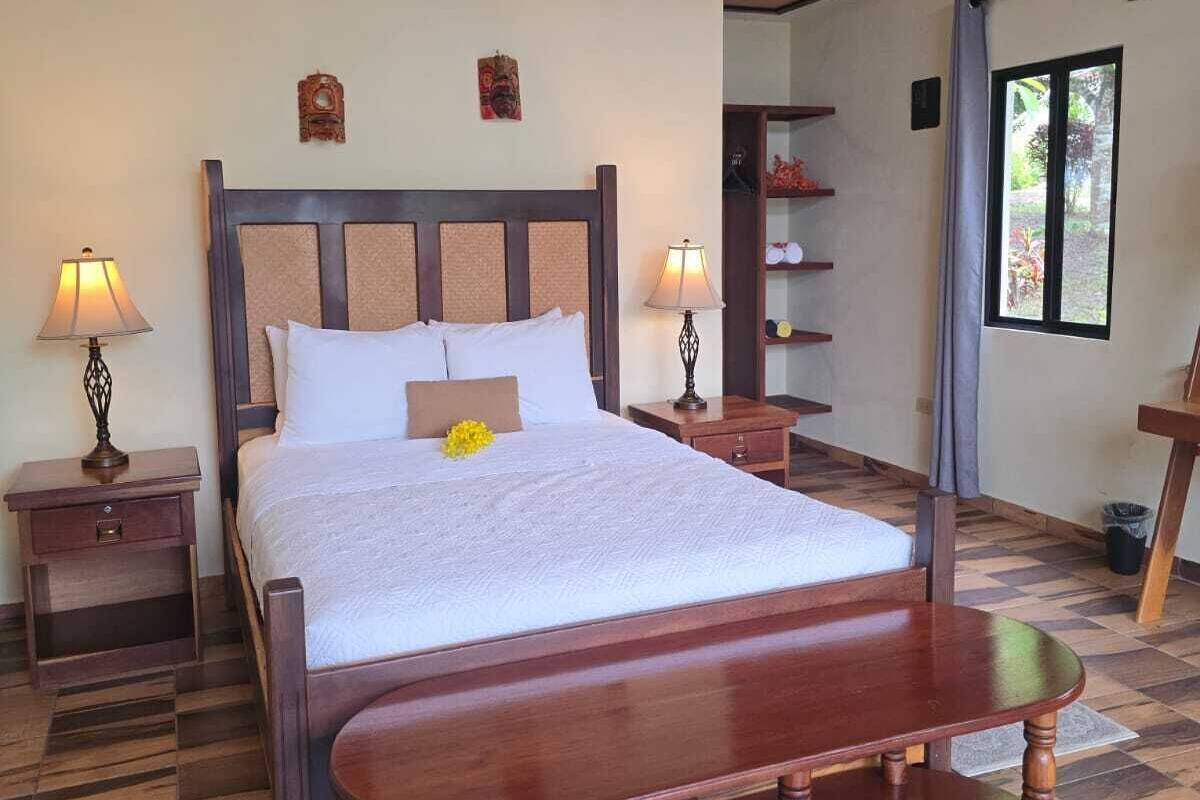 A bedroom with a large bed, wooden headboard, two bedside tables with lamps, a wooden bench, and a view of a garden through a window.