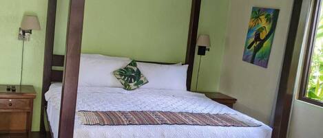 A four-poster bed with a wooden frame, a white bedspread, and a decorative pillow. There are two bedside tables with lamps, a painting on the wall, and a window with a view of greenery.