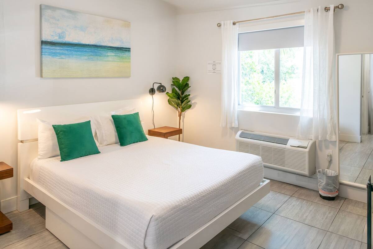 A neatly made bed with white bedding and green pillows, a bedside table with a lamp, a large window with curtains, and a wall-mounted air conditioning unit.