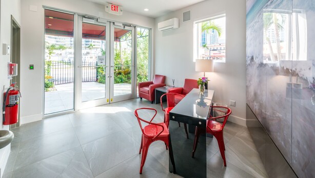 A modern lobby with a glass door entrance, red chairs, a black table, and a wall-mounted air conditioning unit. - Costa Norte Boutique Hotel (Miami Beach)
