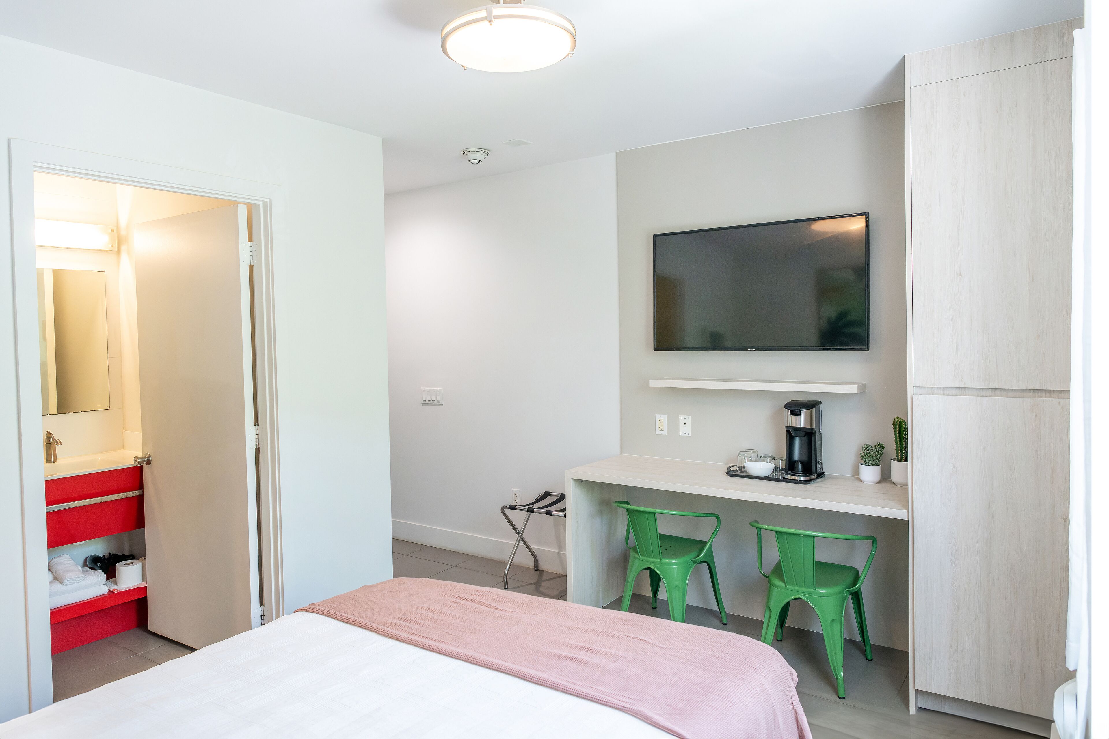 A hotel room with a bed, a desk with two green chairs, a TV, and a red storage unit.