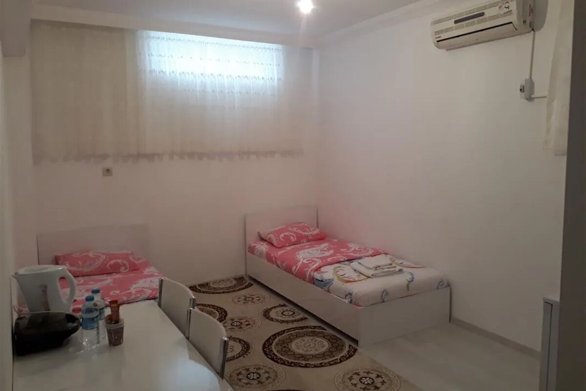 A small, clean room with two single beds, a desk with a chair, a water bottle, a kettle, and a wall-mounted air conditioner.