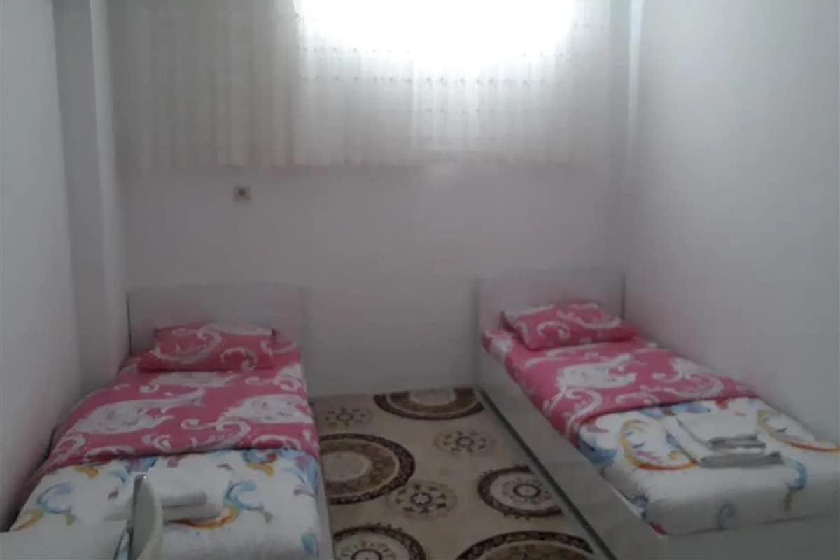 A small, single-room with two beds, a window with sheer curtains, and a patterned floor.