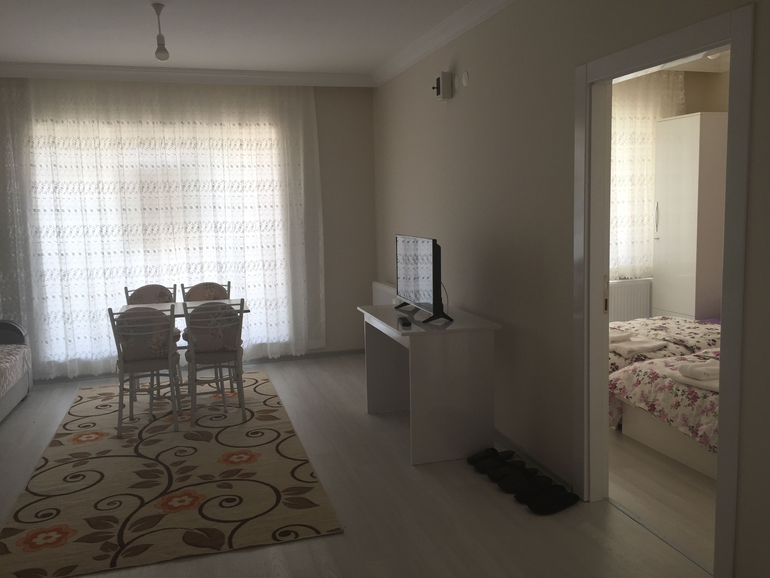 Quarto com uma mesa de jantar, cadeiras, televisão, cama e porta que dá para outro cômodo.