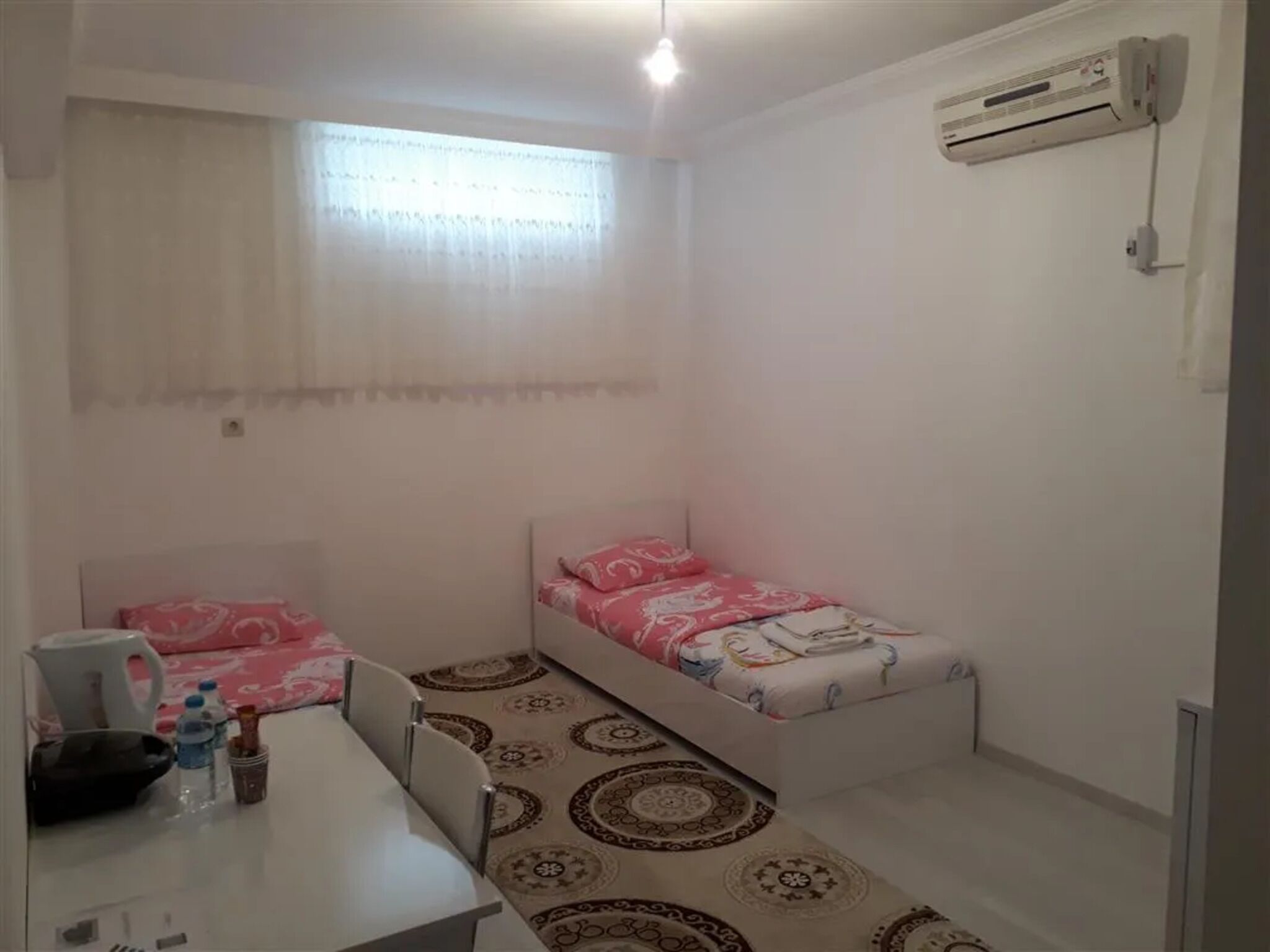 Quarto pequeno e limpo com duas camas individuais, uma escrivaninha com cadeira, garrafa de água, chaleira e ar-condicionado fixado na parede.
