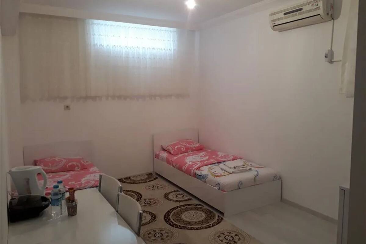 A small, clean room with two single beds, a desk with a chair, a water bottle, a kettle, and a wall-mounted air conditioner.