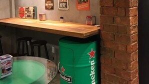 Un espace bar avec un comptoir en bois, des tabourets noirs et un fût vert portant l’inscription « Heineken ».