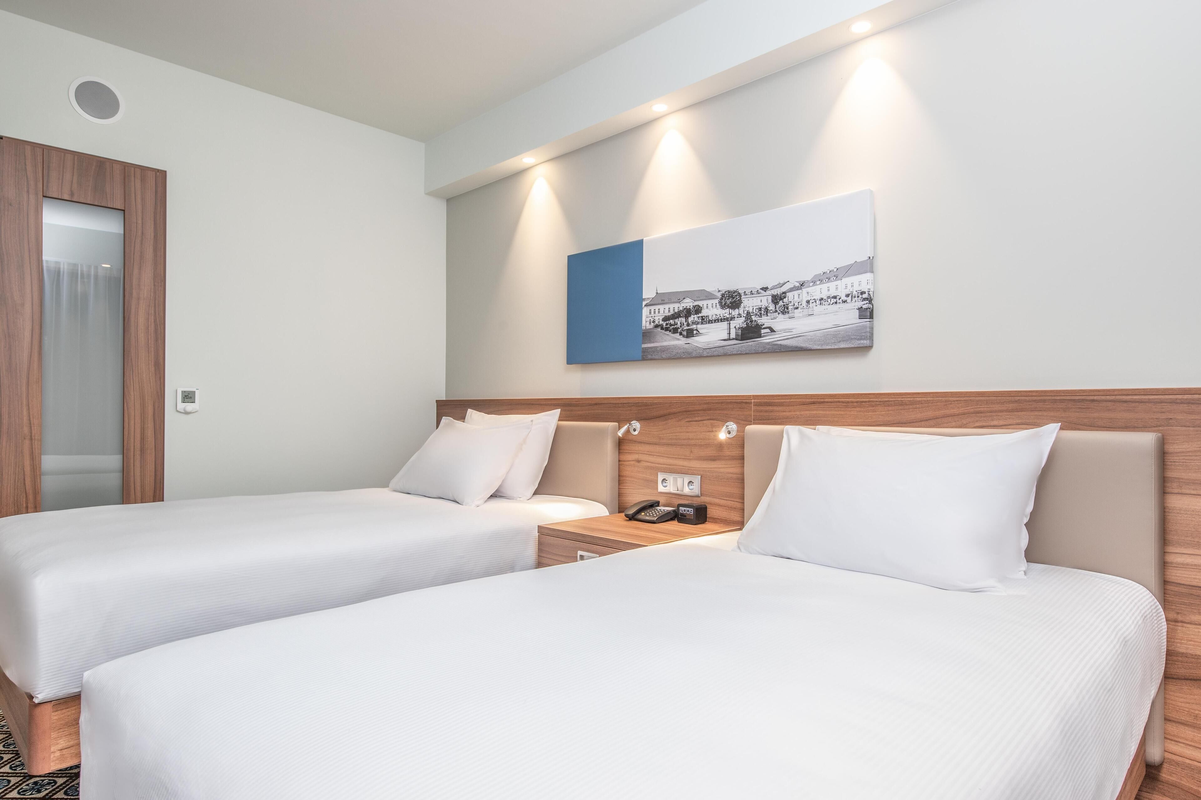 Quarto de hotel com duas camas, uma cabeceira de madeira, um criado-mudo com telefone e um quadro pendurado na parede.