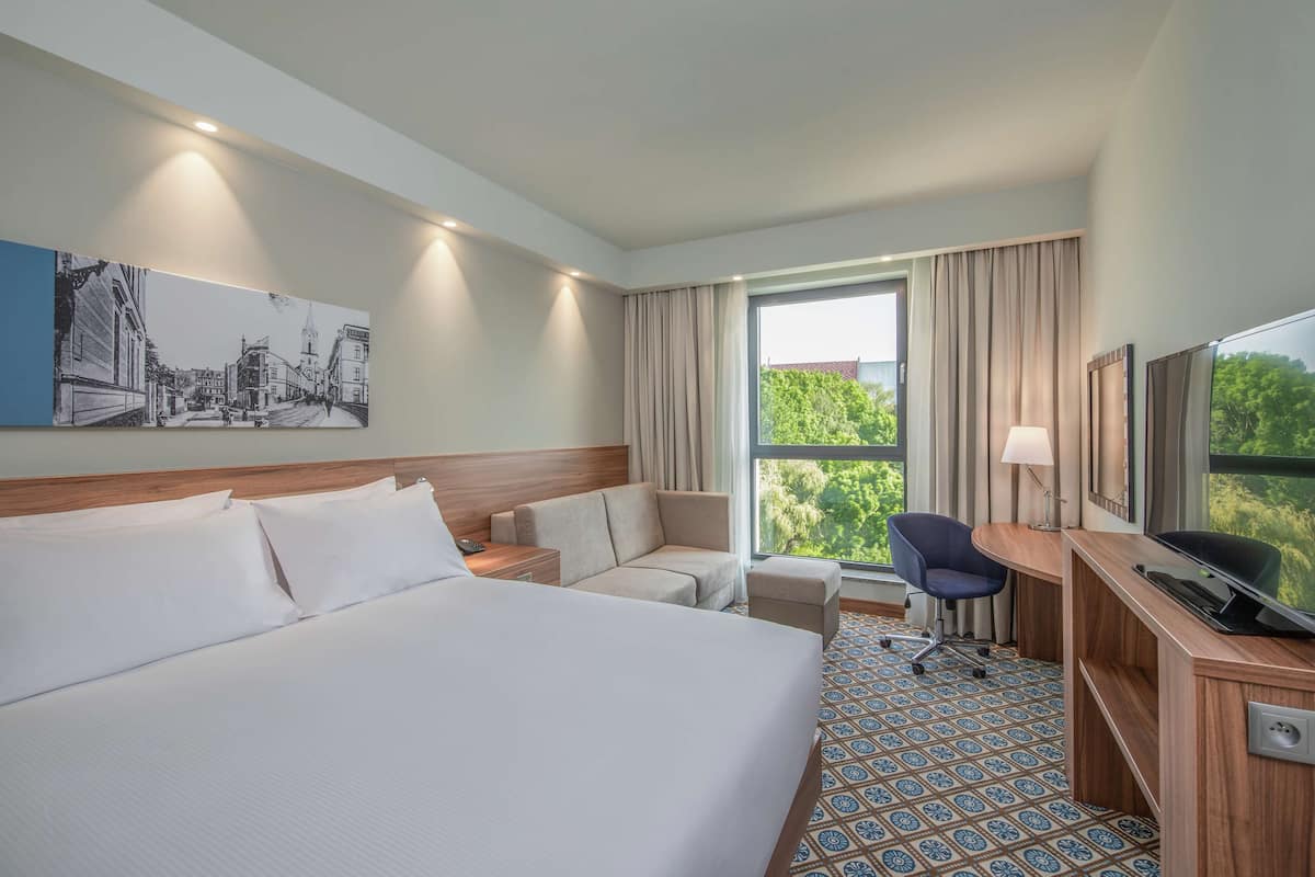 Ein Hotelzimmer mit einem großen Bett, einem Schreibtisch, einem Sessel, einem Fernseher und einem Fenster mit Vorhängen.