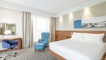 A hotel room with a large bed, a desk with a chair, a small armchair, and a large window with curtains.