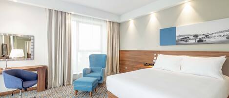 A hotel room with a large bed, a desk with a chair, a small armchair, and a large window with curtains.