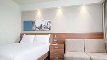 Habitación de hotel con cama, mesita de noche con cajones, sofá, obra de arte en la pared y suelo estampado.