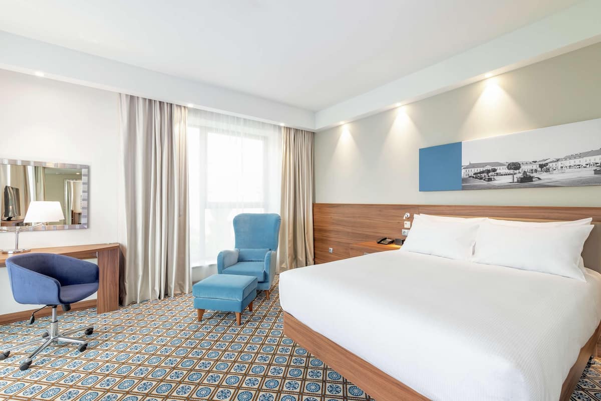 Ein Hotelzimmer mit einem großen Bett, einem Schreibtisch mit Stuhl, einer kleinen Couch und einem großen Fenster mit Vorhängen.