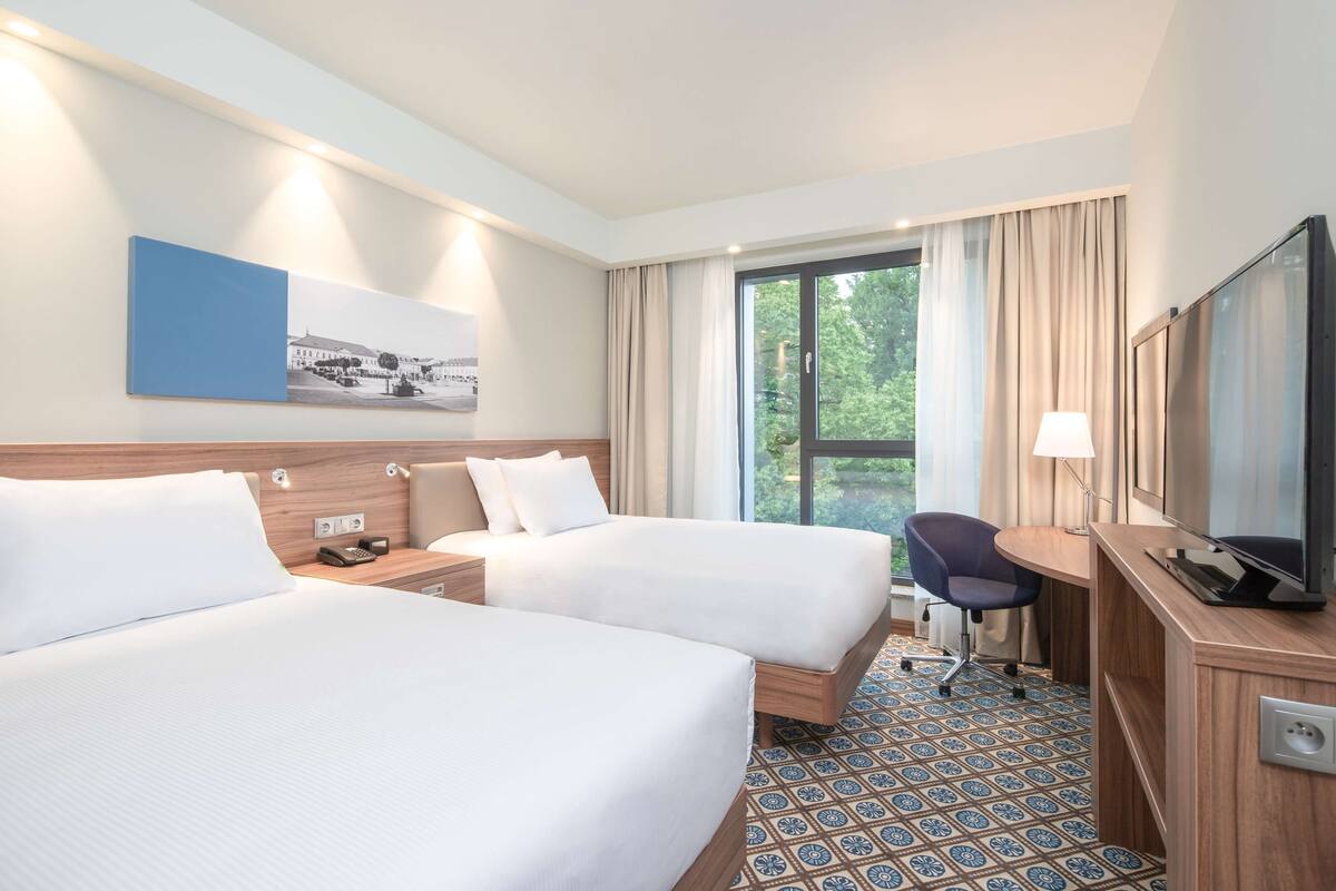 Ein Hotelzimmer mit zwei Betten, einem Schreibtisch, einem Stuhl, einem Fernseher und einem Fenster mit Vorhängen.